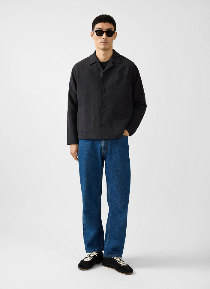 A full length shot of a model wearing the Percival black linen overshirt, blue jeans, sunglasses and black trainers.