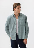 A man models the Percival Clancy Striped Long Sleeve Shirt in blue, worn open over a white vest and dark jeans.