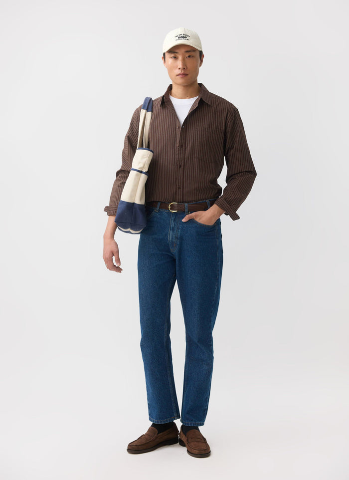 A model wears the brown striped Clancy shirt by Percival, styled with blue jeans, a tote bag, and a baseball cap.