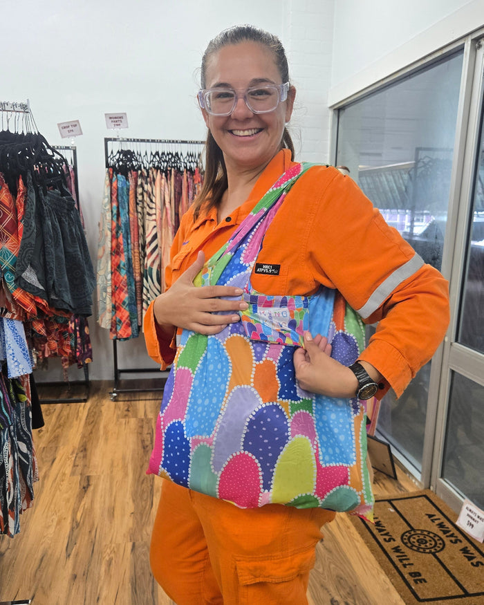 Person wearing an orange safety jacket holding a colorful Aboriginal patterned shopping bag in a store setting.