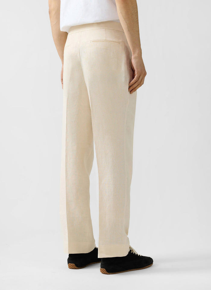 Percival cream linen pleated tailored trousers, shown from the rear and styled with a white t-shirt and black trainers.