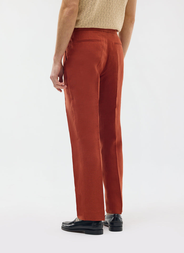 A model from behind wearing the Percival Pleated Tailored Trousers in rust linen with black loafers.