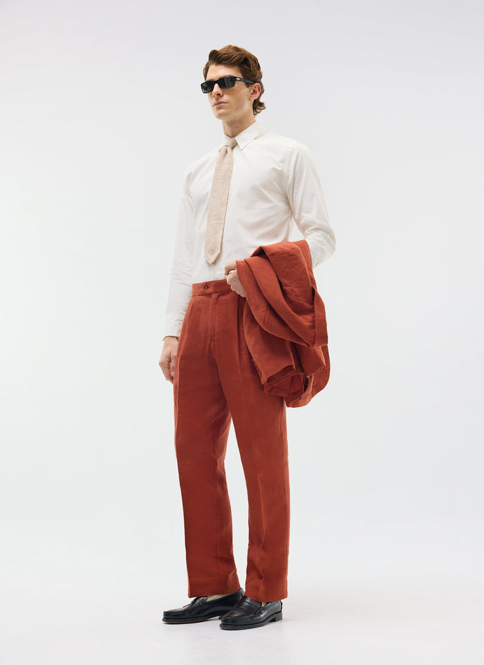 A model wears Percival rust linen pleated tailored trousers with a white shirt and tie, holding the matching jacket.