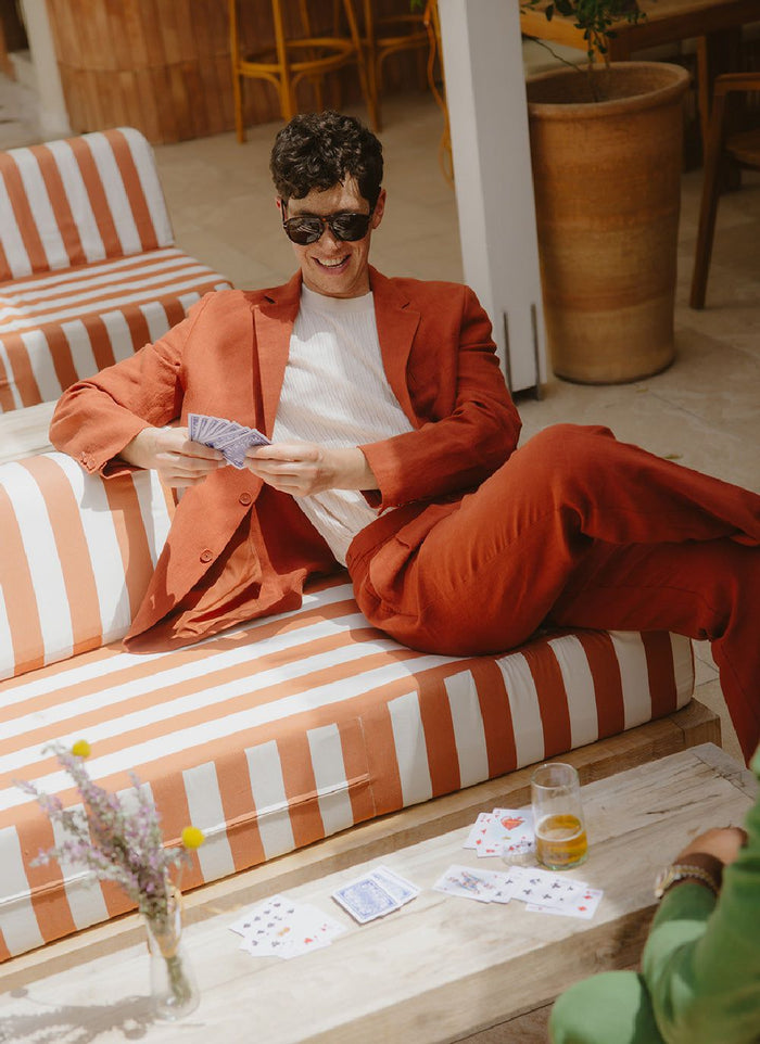A man wearing the Percival rust linen pleated tailored trousers and matching blazer, smiling as he plays cards on a patio.