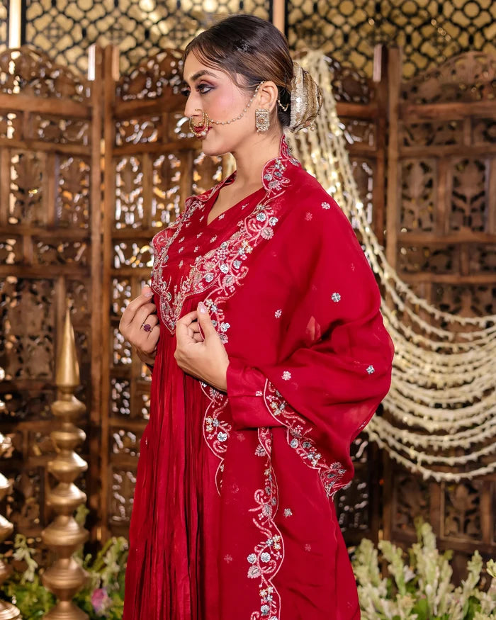 Woman in a red embroidered traditional outfit with intricate designs, standing against an ornate wooden and decorative background.