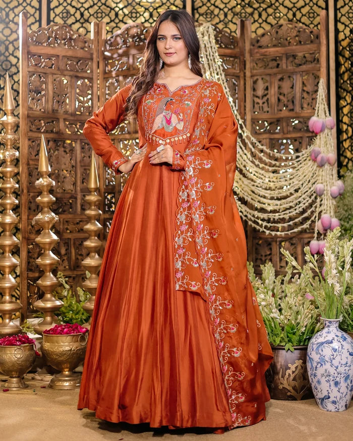 Woman in an orange embroidered traditional outfit standing in a decorative setting with plants and decor.