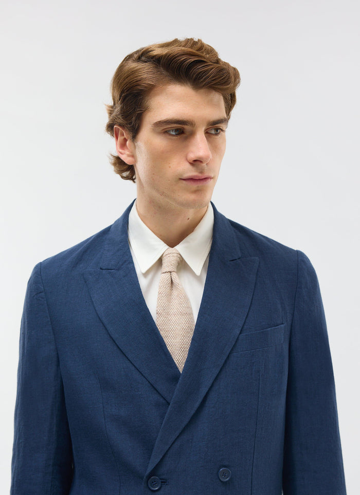 close up of a man wearing the Percival navy double breasted linen blazer with a white shirt and beige knitted tie