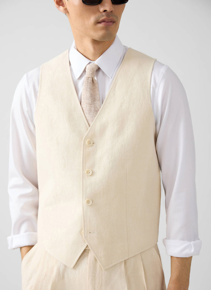A model wears the Percival cream linen waistcoat, styled with a white shirt, textured tie, and matching linen trousers.